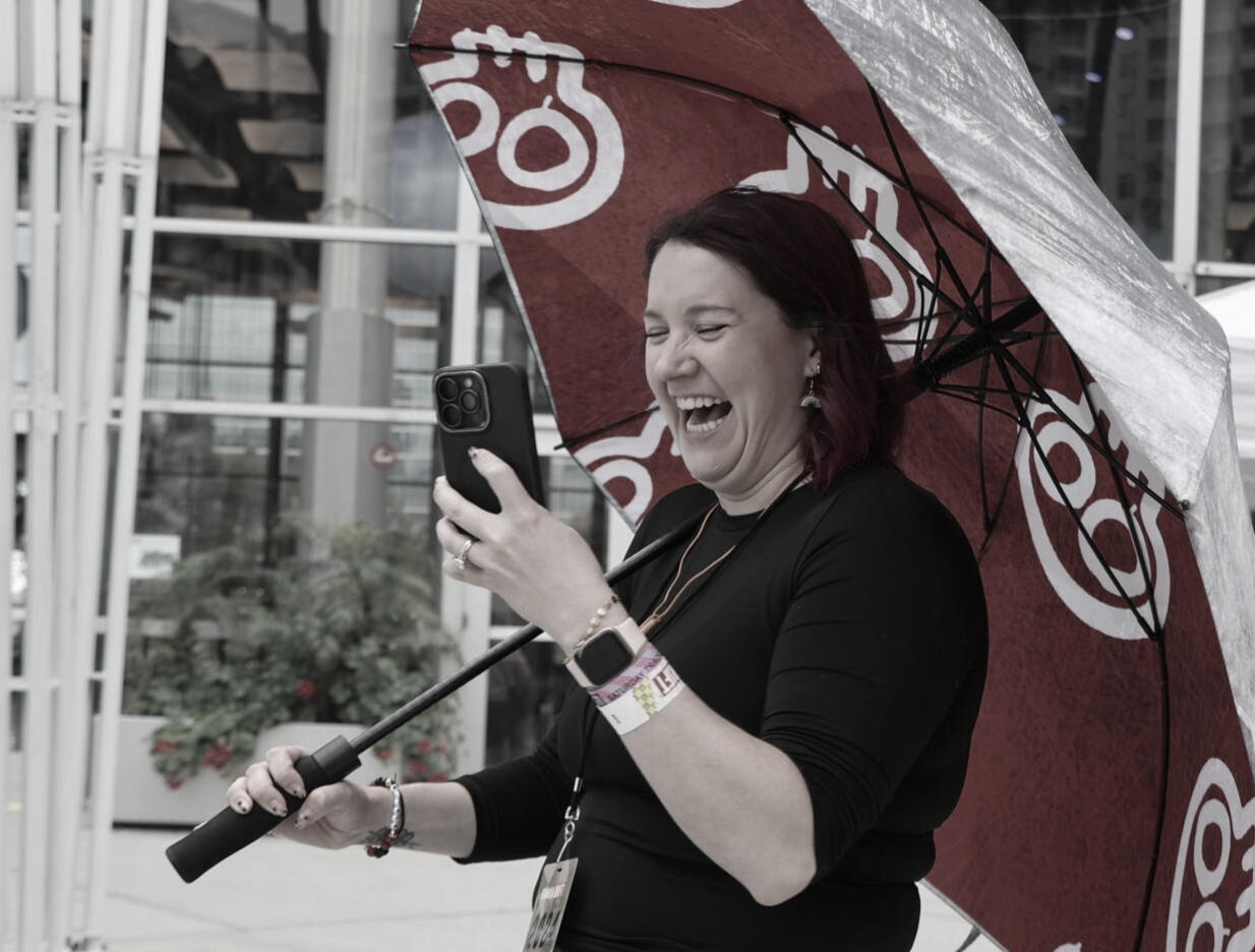Nicole trackside filming content on her phone and laughing while holding an umbrella over herself.